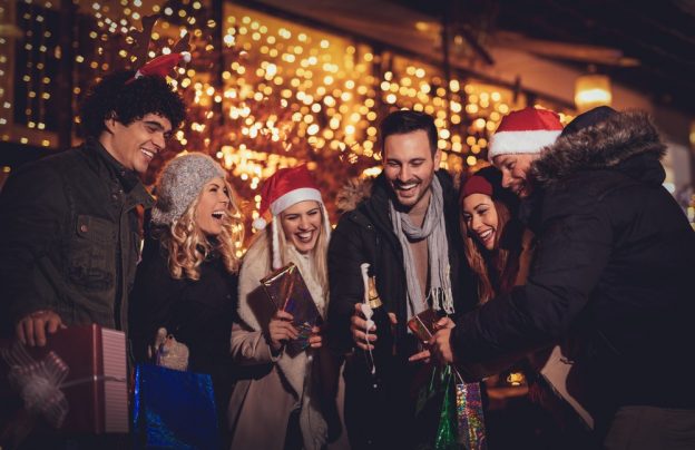 Friends laughing and holding drinks and presents at an outdoor holiday party.